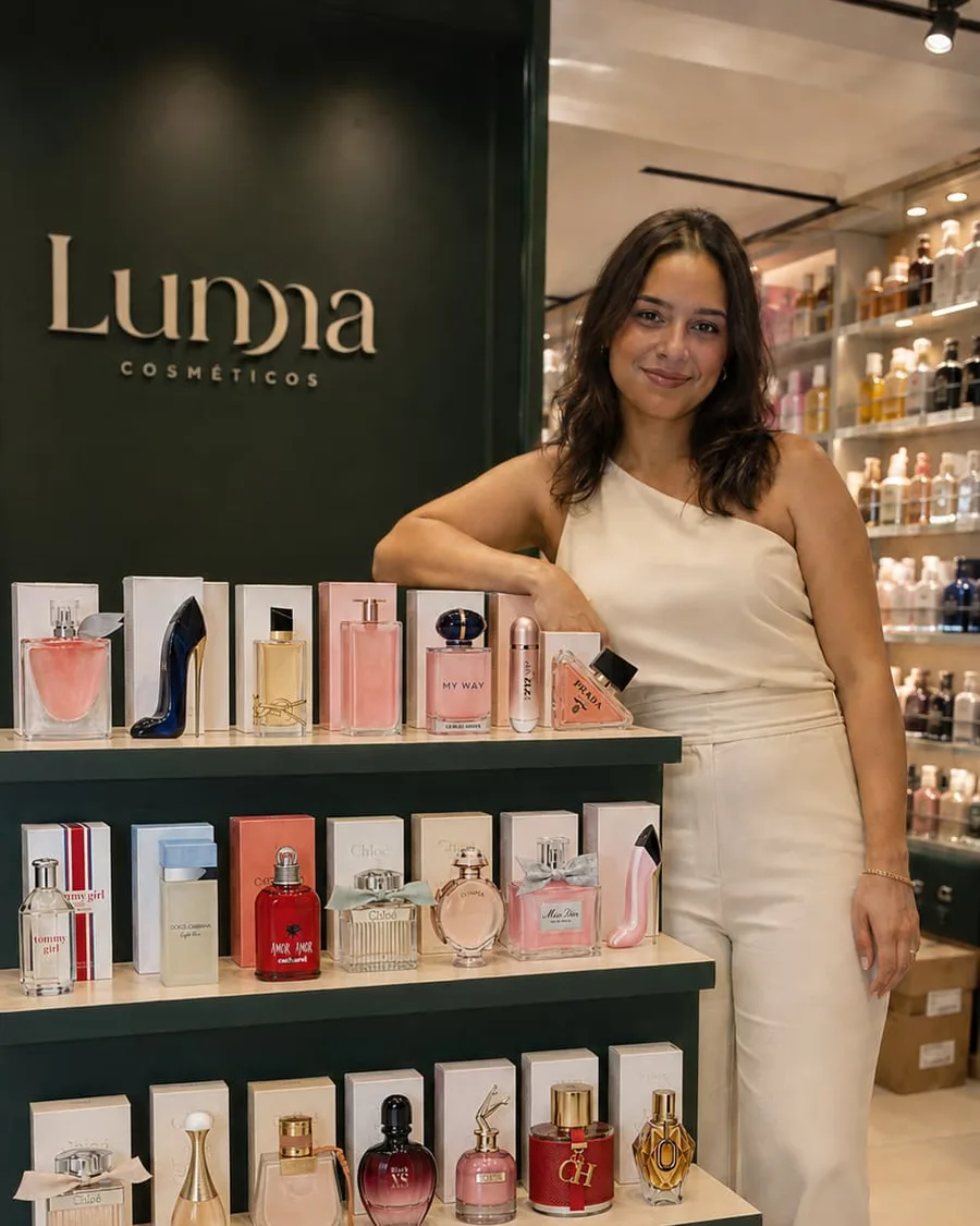 Atendente da Lunna Cosméticos ao lado da vitrine de perfumes da loja, com a marca impressa na parede